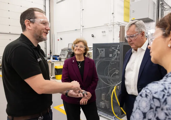 A man in a black polo shirt demonstrates something to three people wearing safety glasses in an industrial setting