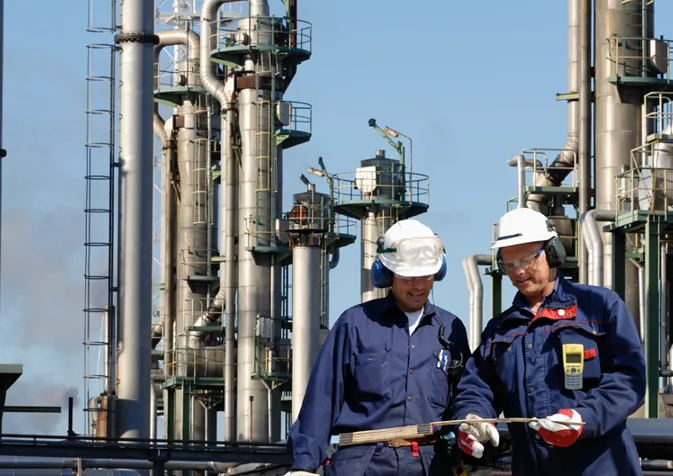 Two men in blue overalls and hard hats standing in front of a refinery