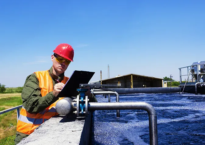 A man in an orange vest and hard hat writing using a clipboard beside an open tank