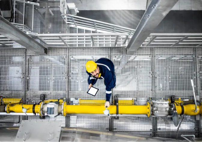 A person in a hard hat and safety glasses stands on a metal walkway with a hand on a yellow pipe.