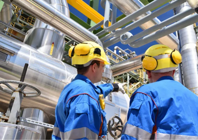 Two people in blue overalls and yellow helmets are working on a large industrial pipe