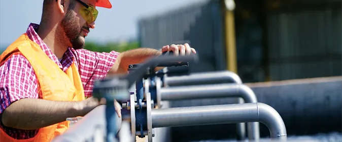 A man in an orange vest and safety glasses is standing on a metal pipe